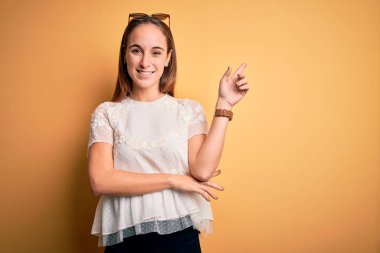 İzole edilmiş sarı arka plan üzerinde günlük tişört ve güneş gözlüğü takan genç ve güzel bir kadın. Yüzünde kocaman bir gülümseme, eli ve parmağı kameraya bakıyor..