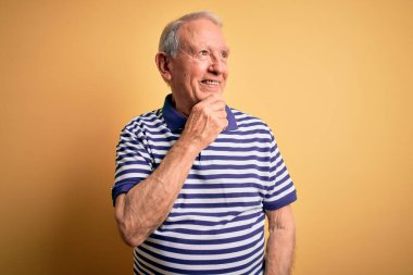 Gri saçlı, donanma çizgili tişört giyen, sarı arka planda duran, elleri çenesinde soru soran, düşünceli bir adam. Düşünceli bir yüzle gülümsüyor. Şüphe kavramı.