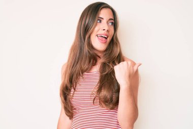 Güzel, genç, beyaz bir kadın, günlük kıyafetler giyiyor, gülümsüyor, gülümsüyor ve başparmağıyla yan tarafa bakıyor.. 