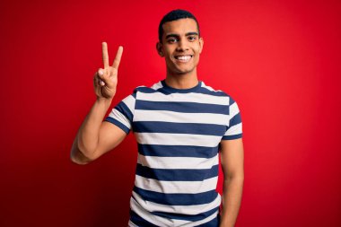 Yakışıklı Afro-Amerikalı sıradan çizgili tişört giyen kırmızı arka planda durup parmaklarını zafer işareti yaparken gösteren kameraya gülümseyen adam. İki numara..