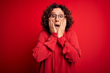 Orta yaşlı güzel kıvırcık saçlı kadın kırmızı arka planda günlük gömlek ve gözlük takıyor. Korkmuş ve şok olmuş, şaşırmış ve şaşırmış bir yüz ifadesi var.