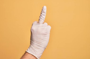 Hand of caucasian young man with medical glove over isolated yellow background showing provocative and rude gesture doing fuck you symbol with middle finger