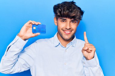 Elinde bir fikir ya da soru işareti ile gülümseyen bir kredi kartı tutan genç İspanyol adam, bir numara. 