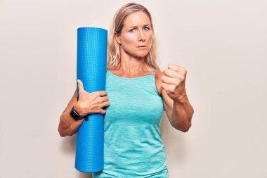 Orta yaşlı, beyaz, elinde yoga minderi tutan sarışın kadın öfkeyle bağırıyor, öfkeyle bağırıyor ve el kaldırıyor. 