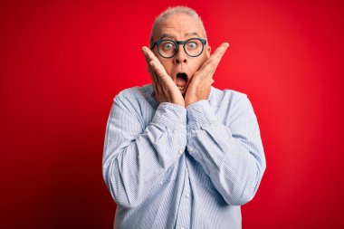 Orta yaşlı, yakışıklı, beyaz bir adam kırmızı arka planda günlük çizgili gömlek ve gözlük takıyor. Korkmuş ve şok olmuş, şaşırmış ve şaşırmış bir yüz ifadesi var.