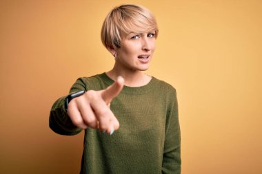 Modern kısa saçlı, sarı arka planda rahat süveter giyen genç sarışın kadın kameraya kızgın ve sinirli olduğunu gösteriyor.