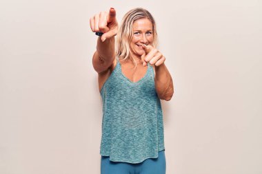 Orta yaşlı beyaz kadın spor kıyafetleri giyiyor. Kamerayı ve seni parmaklarıyla gösteriyor. Gülümsüyor. 