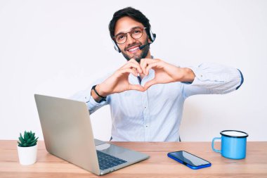 Ofiste çalışan yakışıklı İspanyol adam operatör kulaklığı takıyor. Sevgiyle gülümsüyor. Elleriyle kalp sembolü yapıyor. romantik kavram. 