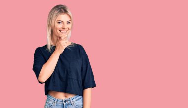 Gündelik tişört giyen genç, sarışın bir kadın kameraya kendinden emin bakıyor kollarını kavuşturmuş ve çenesini kaldırmış bir şekilde gülümsüyor. Olumlu düşünmek. 