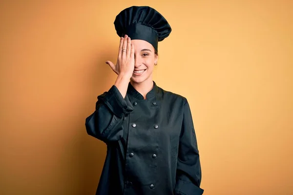 年轻美丽的厨师女士 身穿炊事服 头戴黄底帽 一只手罩着一只眼睛 脸上挂着自信的微笑 感情令人惊讶 — 图库照片