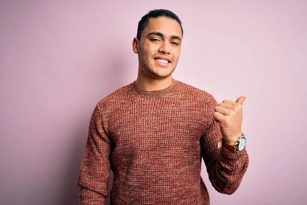 İzole edilmiş pembe arka planda dikilen Brezilyalı genç adam gülümsüyor ve başparmağıyla yan tarafa bakıyor..