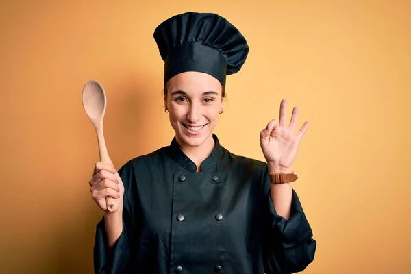 Young beautiful chef woman wearing cooker uniform and hat holding wooden spoon doing ok sign with fingers, excellent symbol