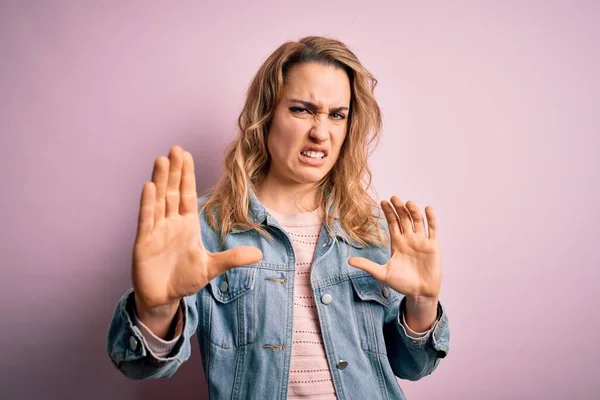 Young beautiful blonde woman wearing casual denim jacket standing over pink background disgusted expression, displeased and fearful doing disgust face because aversion reaction. With hands raised