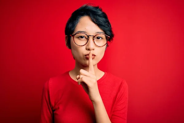 年轻美丽的亚洲女孩穿着休闲的T恤 戴着眼镜 戴着孤立的红色背景 要求保持安静 手指放在嘴唇上 沉默和秘密概念 — 图库照片