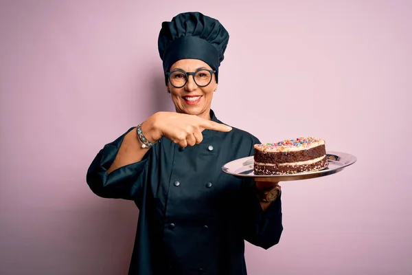 Middle age baker woman wearing cooker uniform and hat holding tray with delicious cake very happy pointing with hand and finger