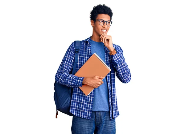 若いアフリカ系アメリカ人の男性学生のバックパックを手であごの質問について考える上で本を保持着て 集中的な表現 笑顔で思いやりのある顔 疑わしい概念 — ストック写真