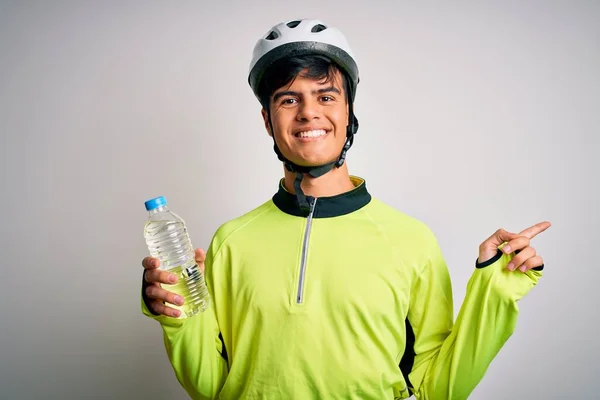 Young handsome cyclist man wearing security bike helmet drinking bottle of water very happy pointing with hand and finger to the side