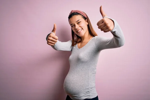 孤立したピンクの背景の上に赤ん坊を期待している若い美しいティーンエイジャーの女の子は手で肯定的なジェスチャーを行うことを承認し 親指を笑顔と成功のために幸せ 勝者のジェスチャー — ストック写真