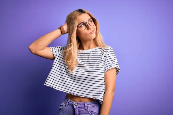 Young beautiful blonde woman wearing striped t-shirt and glasses over purple background confuse and wondering about question. Uncertain with doubt, thinking with hand on head. Pensive concept.