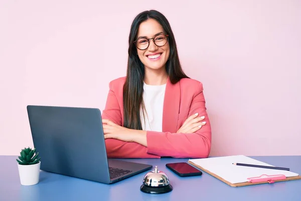 Young caucasian woman sitting at the recepcionist desk working using laptop happy face smiling with crossed arms looking at the camera. positive person.