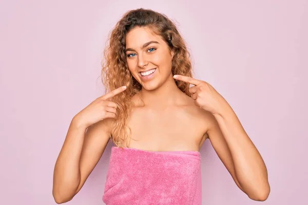 Beautiful blonde woman with blue eyes wearing towel shower after bath over pink background smiling cheerful showing and pointing with fingers teeth and mouth. Dental health concept.