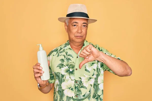 Middle age senior grey-haired man wearing summer hat holding sun protection cream with angry face, negative sign showing dislike with thumbs down, rejection concept