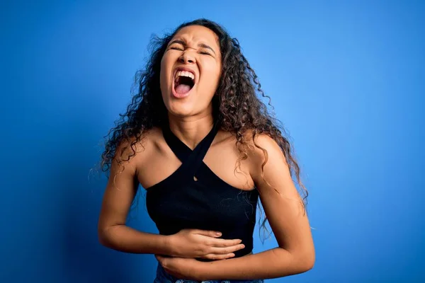 Young beautiful woman with curly hair wearing casual t-shirt standing over blue background with hand on stomach because nausea, painful disease feeling unwell. Ache concept.