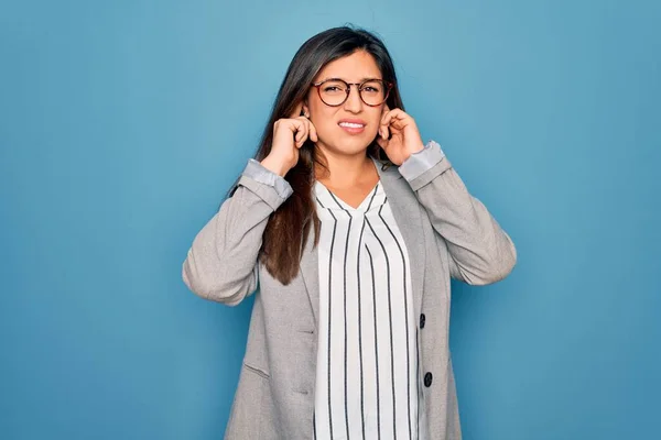 青い隔離された背景の上に立つ眼鏡をかけた若いヒスパニック系のビジネス女性は 大きな音楽のノイズのためにいらいらする表情で指で耳を覆います ろう者の概念 — ストック写真