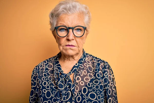 Senior beautiful grey-haired woman wearing casual shirt and glasses over yellow background with serious expression on face. Simple and natural looking at the camera.