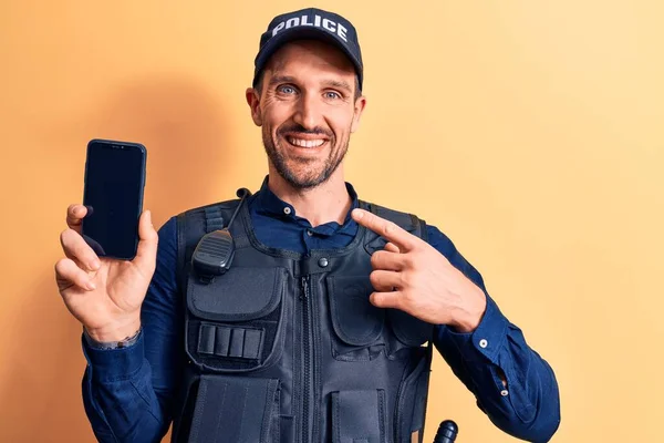 Handsome policeman wearing uniform and bulletprof holding smartphone showing screen smiling happy pointing with hand and finger