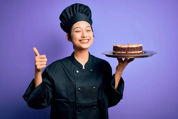中国女厨师身穿炊事服 头戴礼帽 端着蛋糕 笑容满面 做手势 用手指竖起大拇指 神气十足 — 图库照片