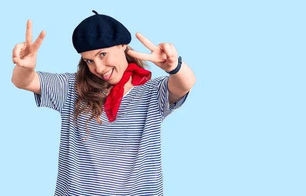 Young beautiful blonde woman wearing french beret and striped t-shirt smiling with tongue out showing fingers of both hands doing victory sign. number two.