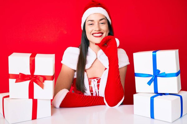 Молодая женщина в костюме Санта-Клауса сидит за столом с подарками, улыбаясь и уверенно глядя в камеру со скрещенными руками и рукой на подбородке. Позитивное мышление. 