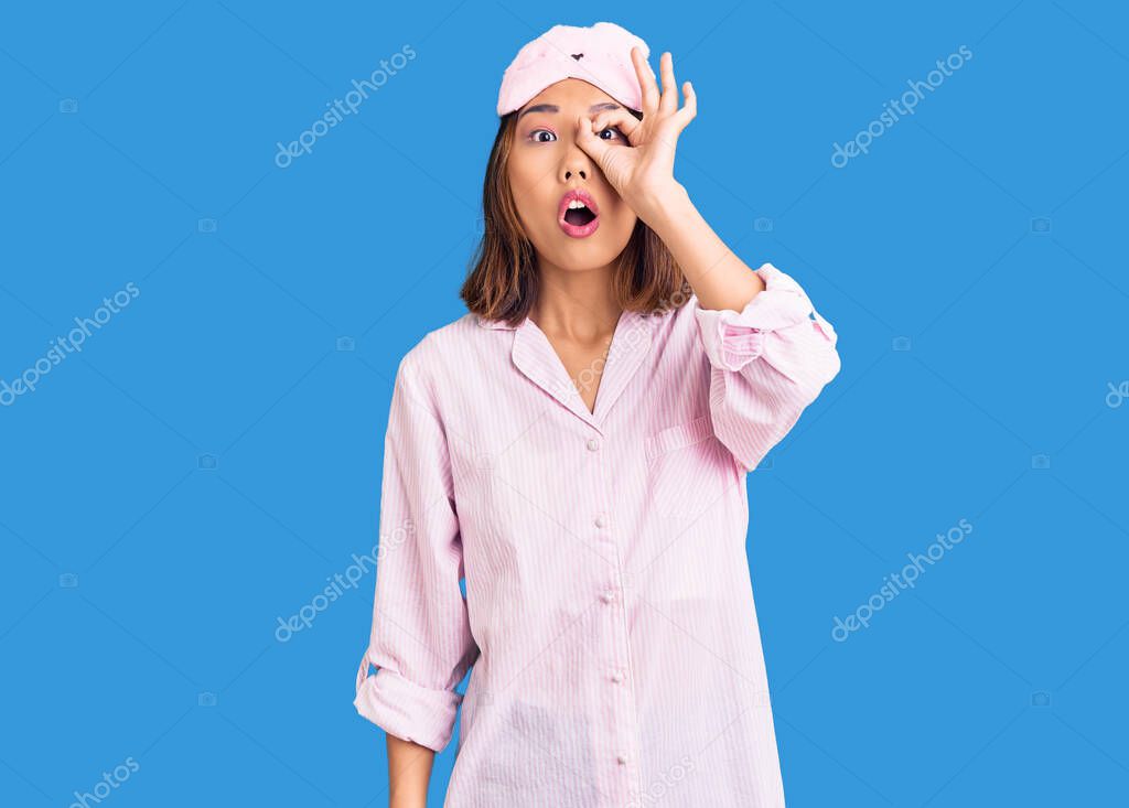 Young beautiful chinese girl wearing sleep mask and pajama doing ok gesture shocked with surprised face, eye looking through fingers. unbelieving expression. 