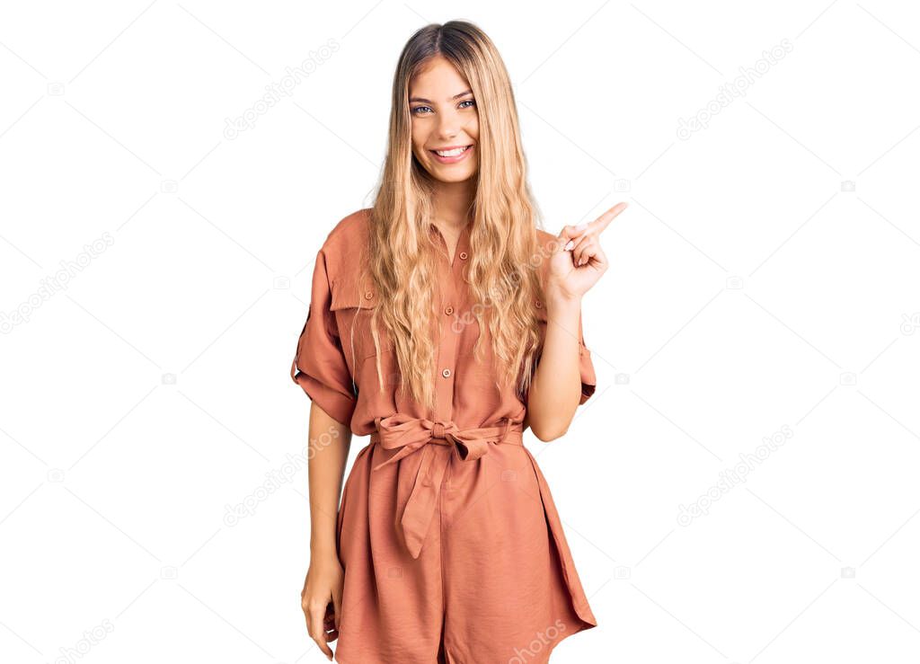 Beautiful caucasian woman with blonde hair wearing summer jumpsuit with a big smile on face, pointing with hand finger to the side looking at the camera. 