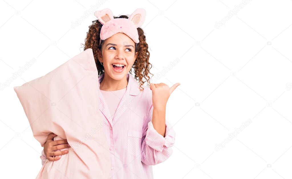 Beautiful kid girl with curly hair wearing sleep mask and pajamas holding pillow pointing thumb up to the side smiling happy with open mouth 