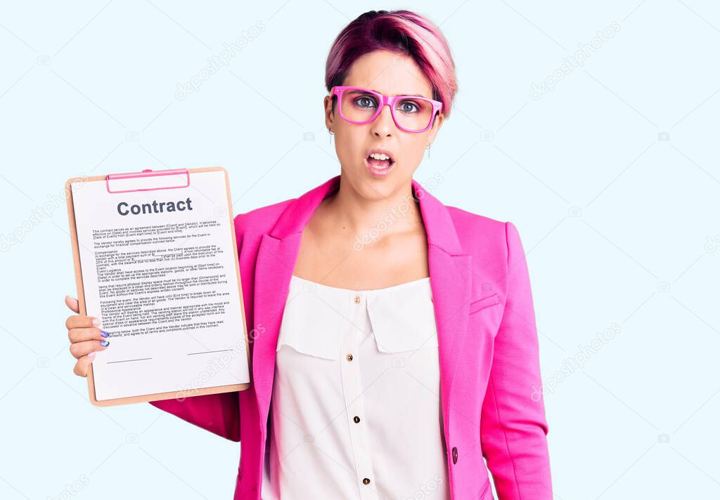 Young beautiful woman with pink hair holding clipboard with contract document in shock face, looking skeptical and sarcastic, surprised with open mouth 