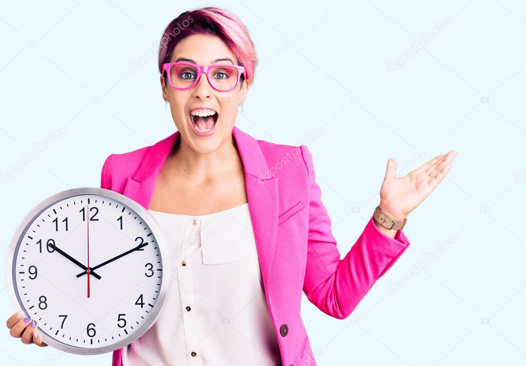 Young beautiful woman with pink hair wearing business jacket and holding clock celebrating victory with happy smile and winner expression with raised hands 