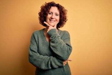 Orta yaşlı, kıvırcık saçlı güzel bir kadın izole edilmiş sarı arka plan üzerine günlük süveter giyip kameraya bakarken kollarını çaprazlayarak ve çenesini kaldırarak gülümsüyor. Olumlu düşünüyorum..