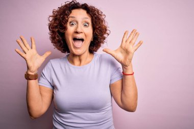 Orta yaşlı güzel kıvırcık saçlı kadın pembe arka plan üzerine gündelik tişört giyip çılgın ve hayret verici başarısını kutlarken kollarını kaldırmış ve gözleri açık bir şekilde heyecanla haykırıyor. Kazanan konsept
