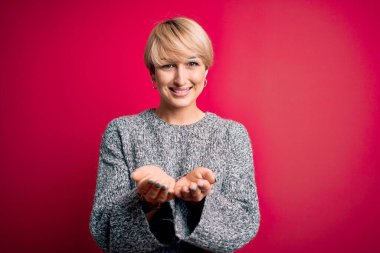 Modern kısa saçlı, pembe arka planda rahat süveter giyen, avuçlarıyla gülümseyen ve el hareketi yapan genç sarışın kadın. Tutun ve koruyun