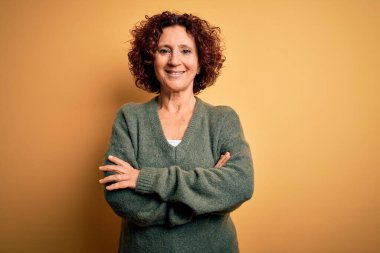 Orta yaşlı güzel kıvırcık saçlı kadın izole edilmiş sarı arka planda rahat süveter giyip kollarını kavuşturup kameraya bakarken gülümsüyor. Pozitif kişi.