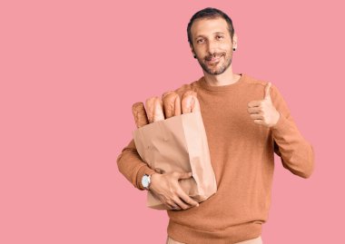 Elinde ekmek dolu kağıt torba tutan genç ve yakışıklı adam mutlu ve olumlu gülümsüyor, başparmağı mükemmel ve onay işareti yapıyor. 