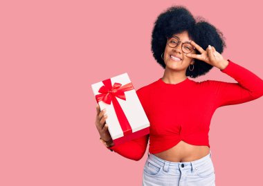 Genç Afrikalı Amerikalı kız elinde hediyeyle barış sembolü yapıyor. Yüzünde parmaklarıyla gülümsüyor ve zaferini gösteriyor. 