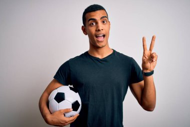 Beyaz arka planda futbol topu tutan yakışıklı Afro-Amerikan adam parmaklarını zafer işareti yaparken gösteren kameraya gülümsüyor. İki numara..
