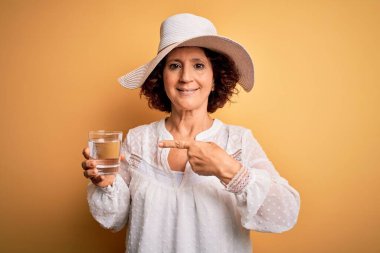 Orta yaşlı kıvırcık kadın tatilde izole edilmiş sarı arka planda bir bardak su içiyor.