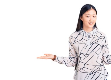 Genç, güzel, Çinli bir kadın, günlük tişört giyiyor, gülümsüyor, avuçlarını açıyor, sunuyor, reklam yapıyor, karşılaştırıyor ve dengede duruyor. 