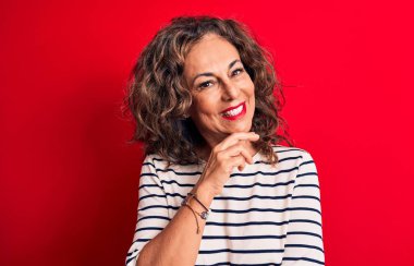Orta yaşlı, esmer, çizgili tişört giyen güzel bir kadın kırmızı arka planda durup gülümsüyor ve kameraya kollarını kavuşturup çenesini kapatıyor. Olumlu düşünüyorum..