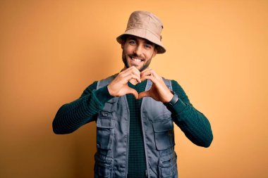 Tatilde sakallı yakışıklı turist sarı arka planda kaşif şapkası takıyor aşkla gülümsüyor elleriyle kalp sembolü yapıyor. Romantik konsept.