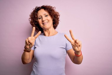 Orta yaşlı güzel kıvırcık saçlı kadın izole edilmiş pembe arka planda günlük tişört giyip parmaklarını zafer işareti yaparken gösteren kameraya gülümsüyor. İki numara..